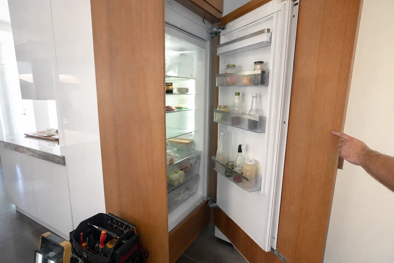 A hand swings open the wood-panelled door of a fully integrated column refrigerator, its lit shelves and door bins showing, a tool bag and work light on the tile floor