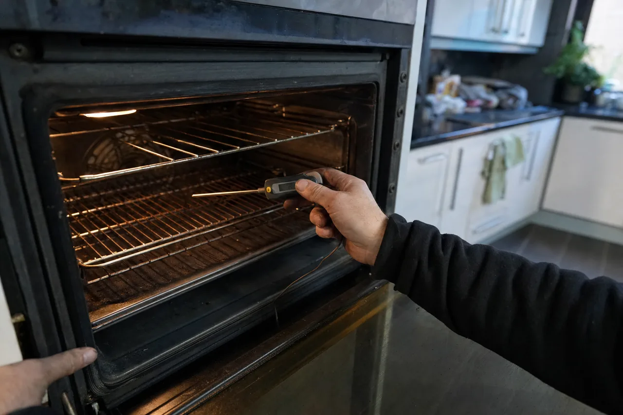 A hand holding a probe thermometer clipped to the middle rack of an open wall oven, the cavity lit and the door held with the other hand