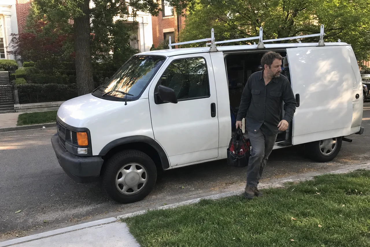 Mirrored view of a white panel van with its side door open and a technician carrying a tool bag toward the sidewalk