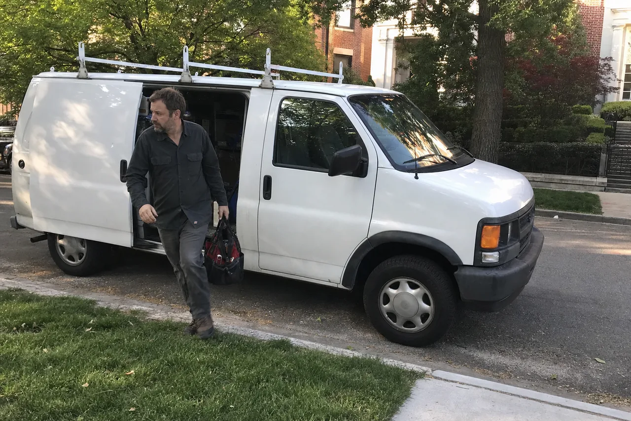 A technician stepping down out of the side door of a white service van parked at the kerb of a shaded residential street, tool bag in his hand and a ladder rack overhead